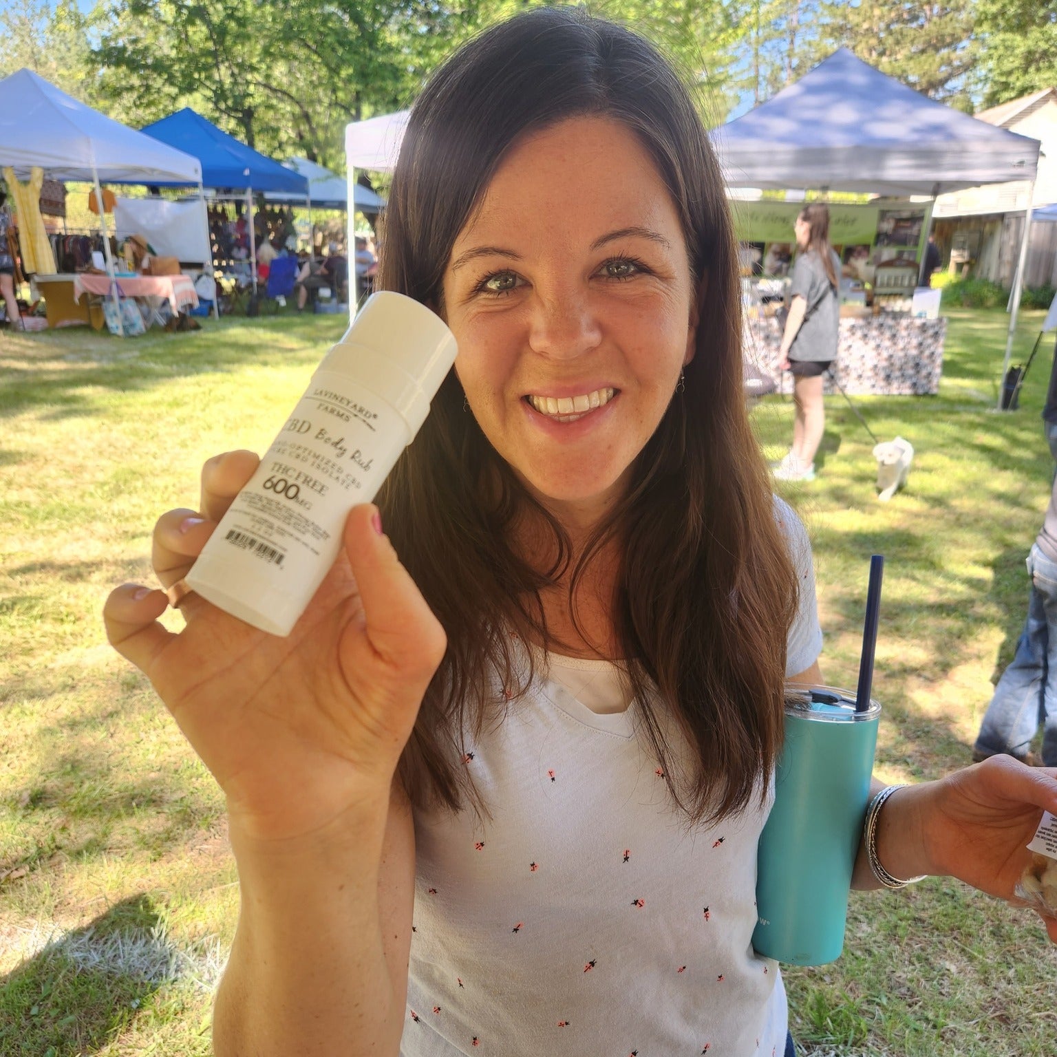 Woman holding a bottle of sunscreen at an outdoor event with tents and people in the background.